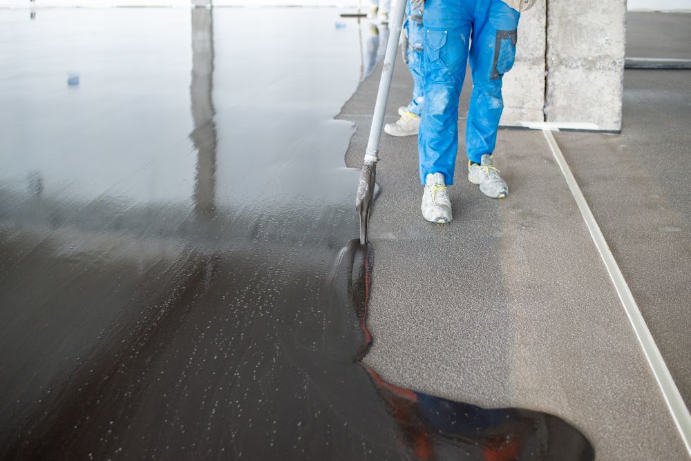 A worker in blue coveralls applies a black liquid sealant or coating to a concrete floor using a long-handled tool, demonstrating expert waterproofing in Singapore.