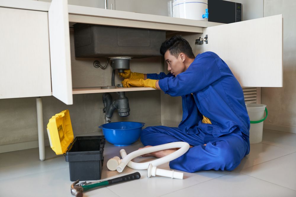 Person wearing blue coveralls and yellow gloves fixing a pipe under a kitchen sink, with tools and parts scattered on the floor nearby—a typical scene during Water Leak Repair: Common Causes and Solutions.